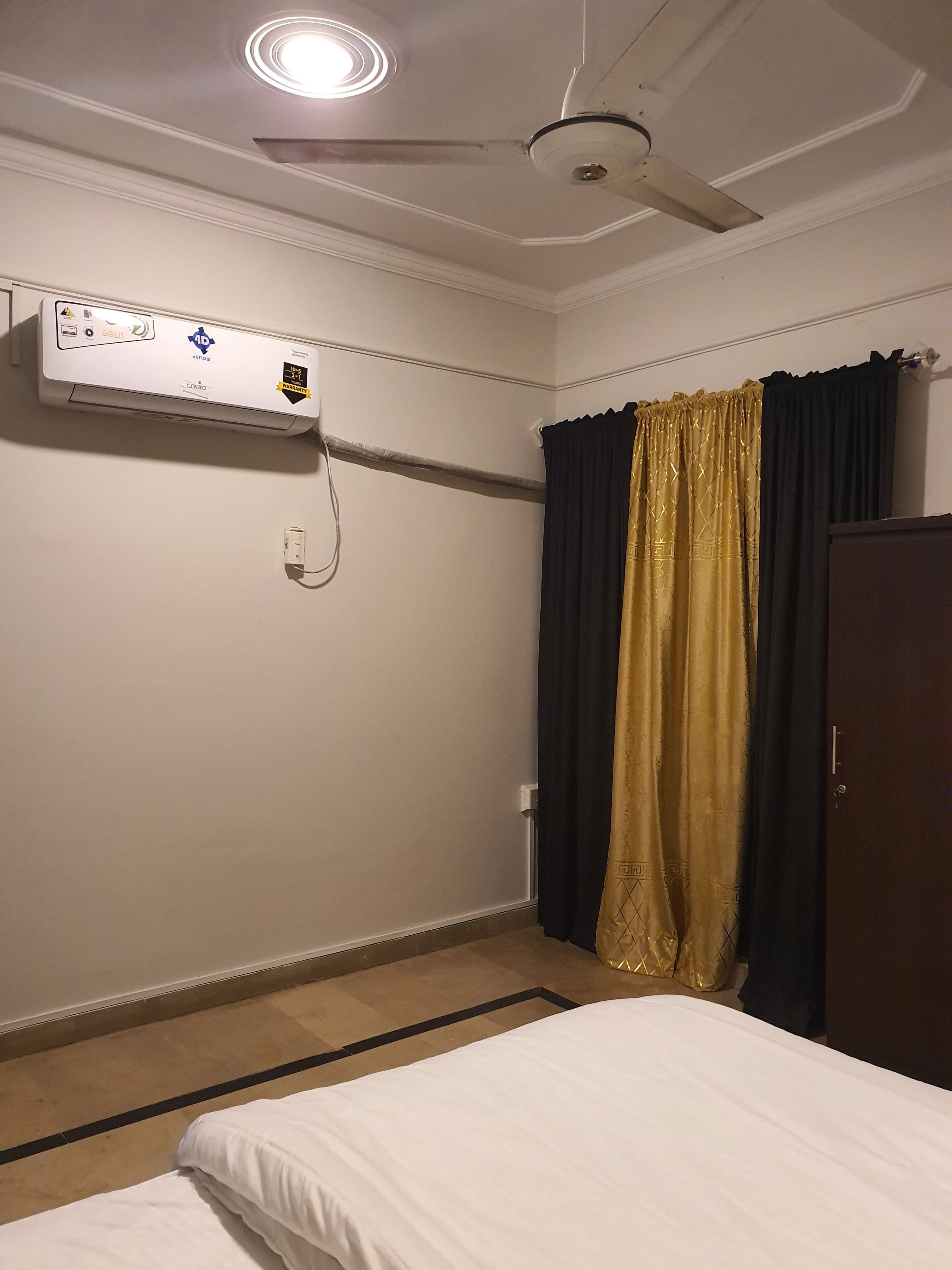 Habitación de hotel con cama, ventilador de techo, aire acondicionado y ventana con cortinas oscuras y doradas.