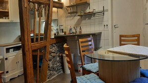 A kitchen with white cabinets, a stone wall, a wooden chair, a glass table, and a blue cushioned chair. - Cosy holiday home near the ski lift (Altenfeld)