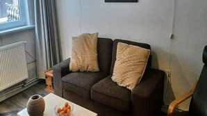 A living room with a dark brown sofa, two beige pillows, a white vase, and a bowl of oranges on a white table. - Cosy holiday home near the ski lift (Altenfeld)