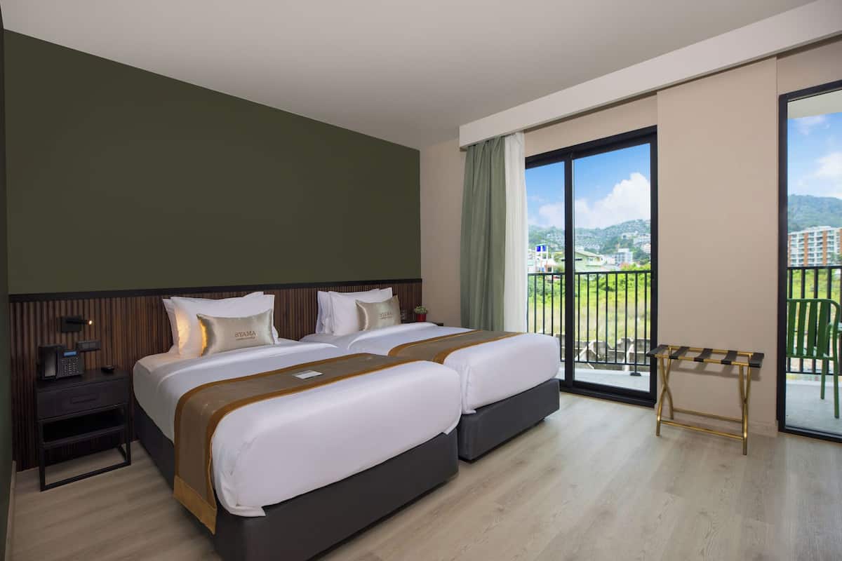 A hotel room with two beds, a nightstand, a small table, and a balcony with a view of buildings and greenery.