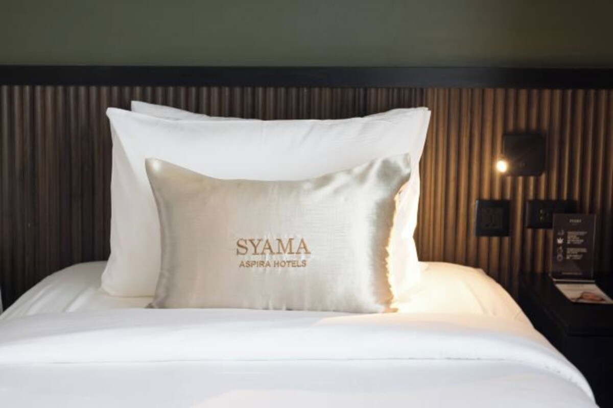 A neatly made bed with white bedding and two pillows, one of which has the text "SYAMA ASPIRA HOTELS" on it.
