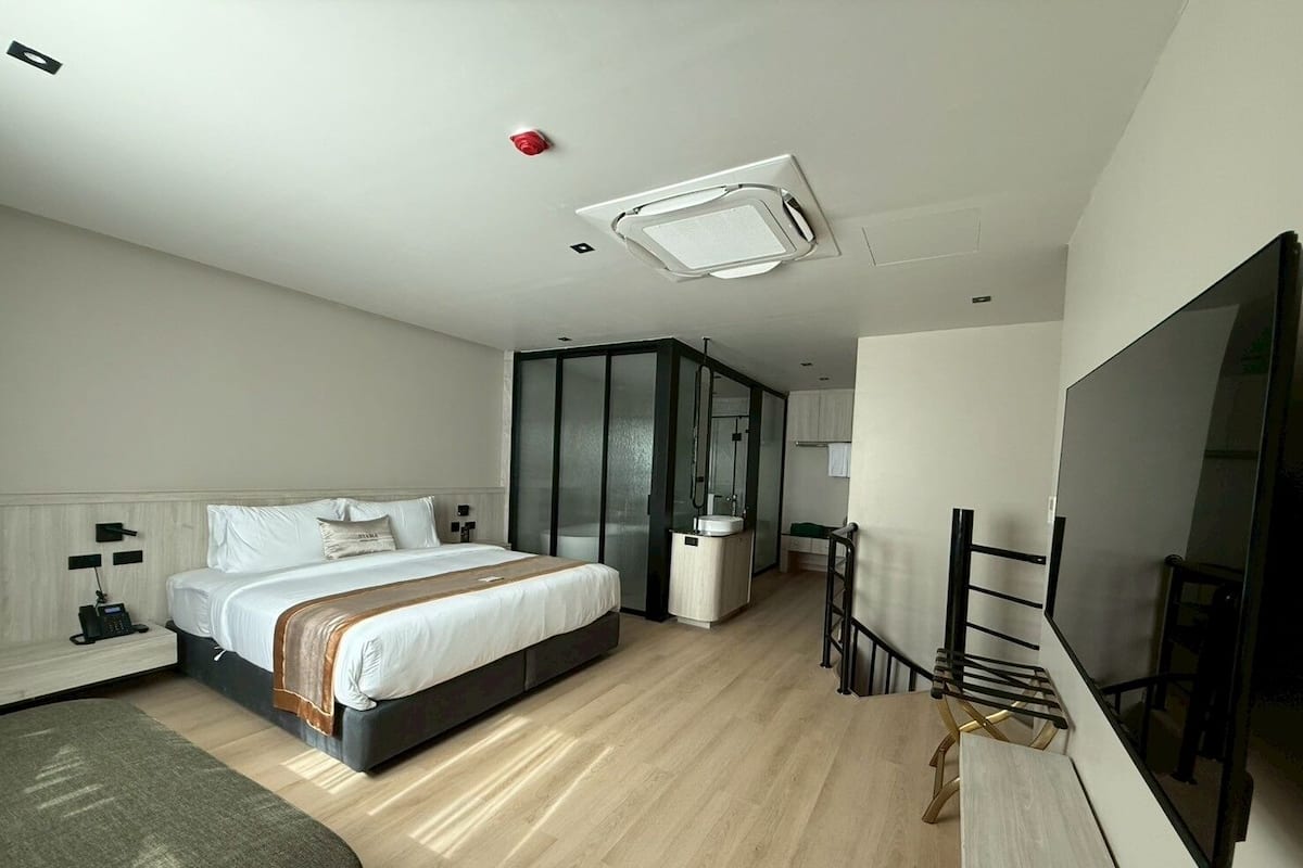 A modern hotel room with a large bed, a desk, a TV, and a bathroom with a glass partition.