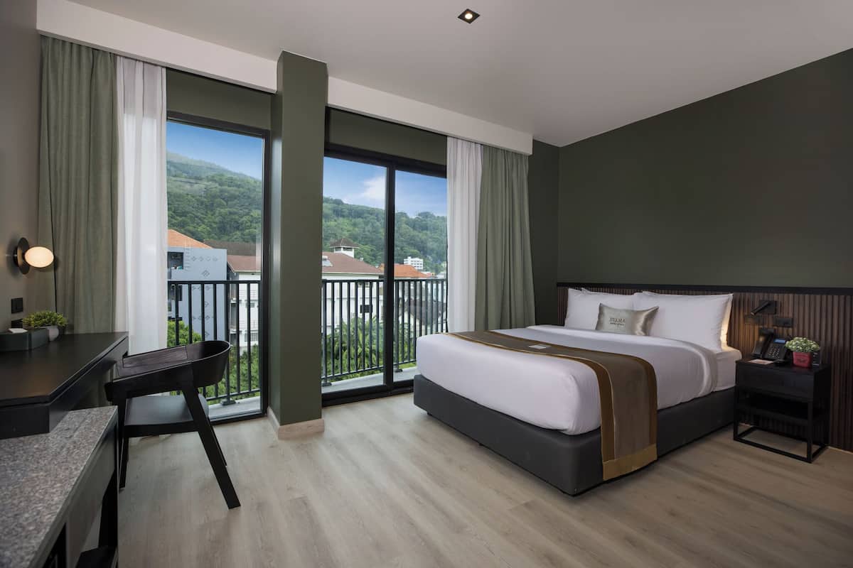 A hotel room with a large bed, a desk, and a view of a balcony with greenery and buildings.