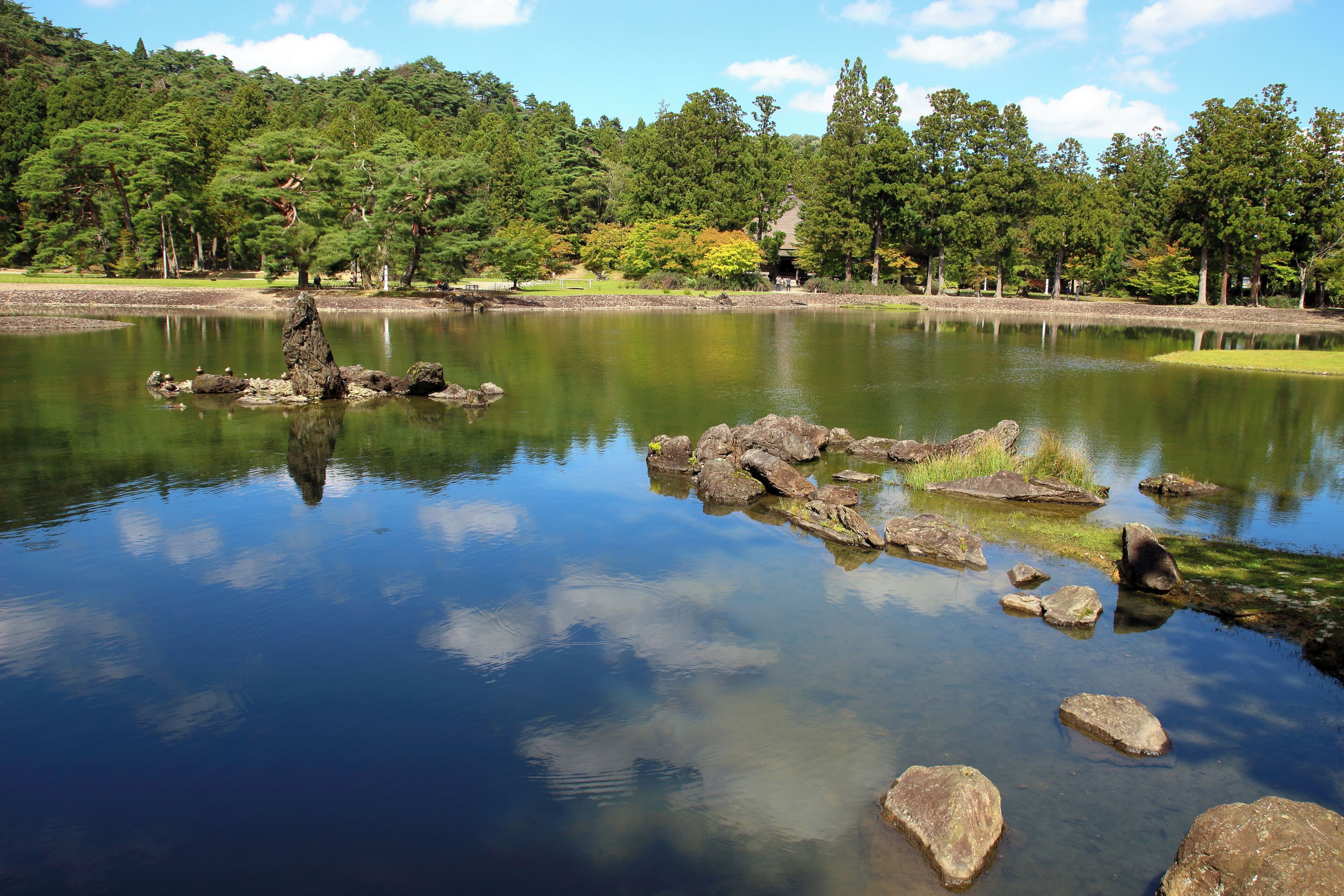 A tranquil lake with clear reflections, surrounded by dense forest and a few structures in the background.