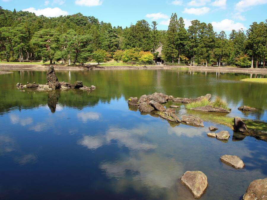 A tranquil lake with clear reflections, surrounded by dense forest and a few structures in the background.