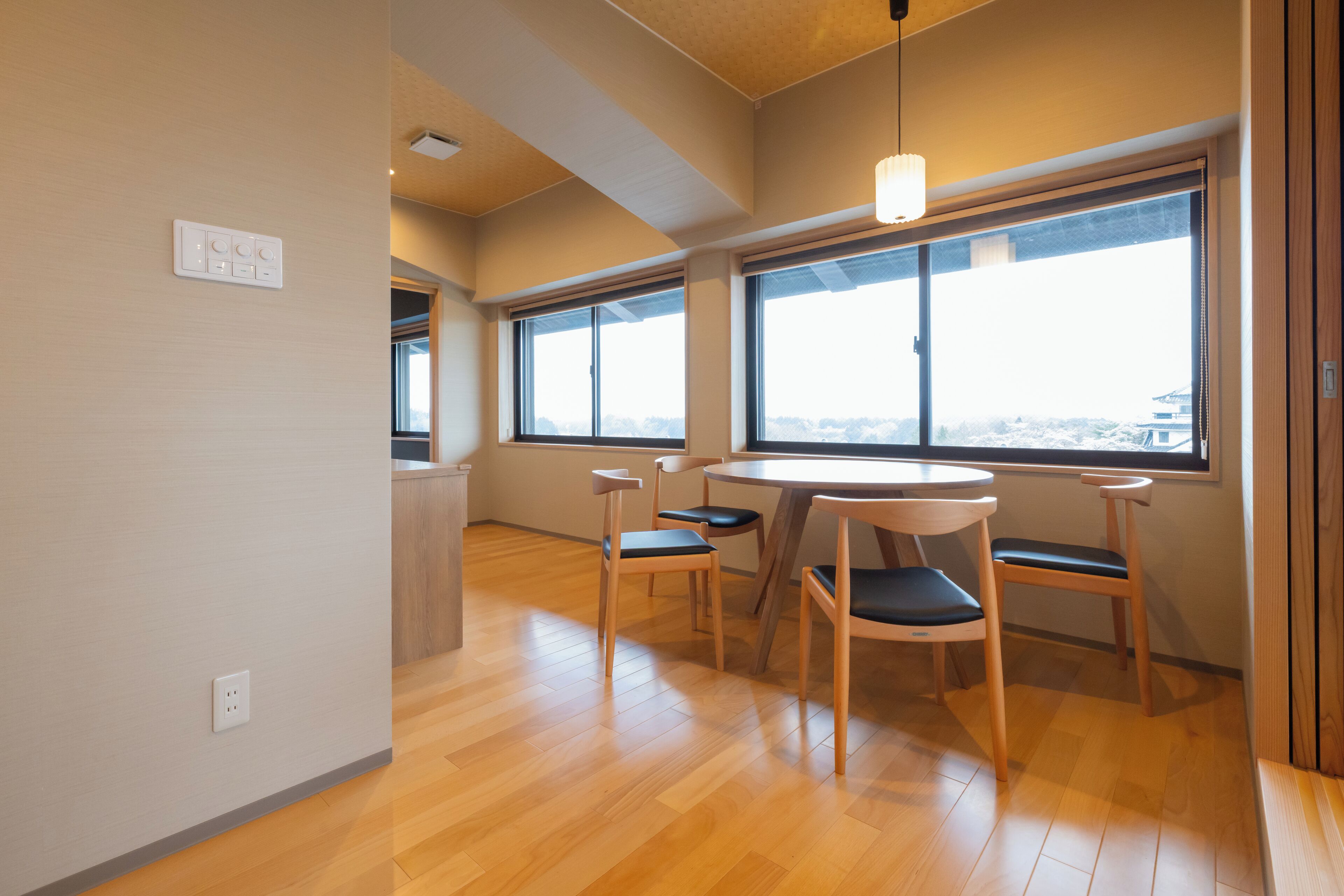 A modern room with a wooden floor, a round table with four chairs, large windows with a view, and a pendant light.