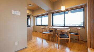 A modern room with a wooden floor, a round table with four chairs, large windows with a view, and a pendant light.