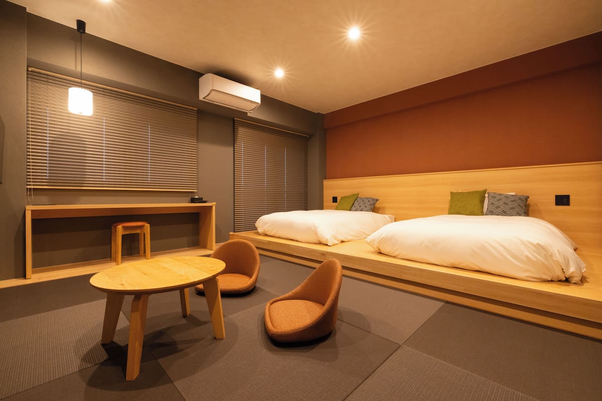Une chambre d’hôtel moderne avec un lit en bois, deux chaises, une petite table et une grande fenêtre avec des stores.