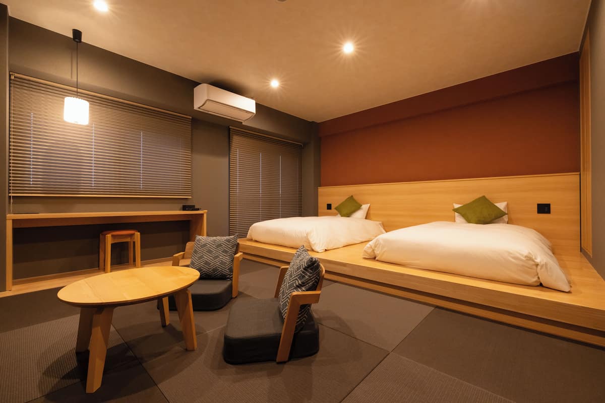 Une chambre d’hôtel comprenant un lit, deux tables de chevet, une petite table, deux chaises et une fenêtre avec des stores.