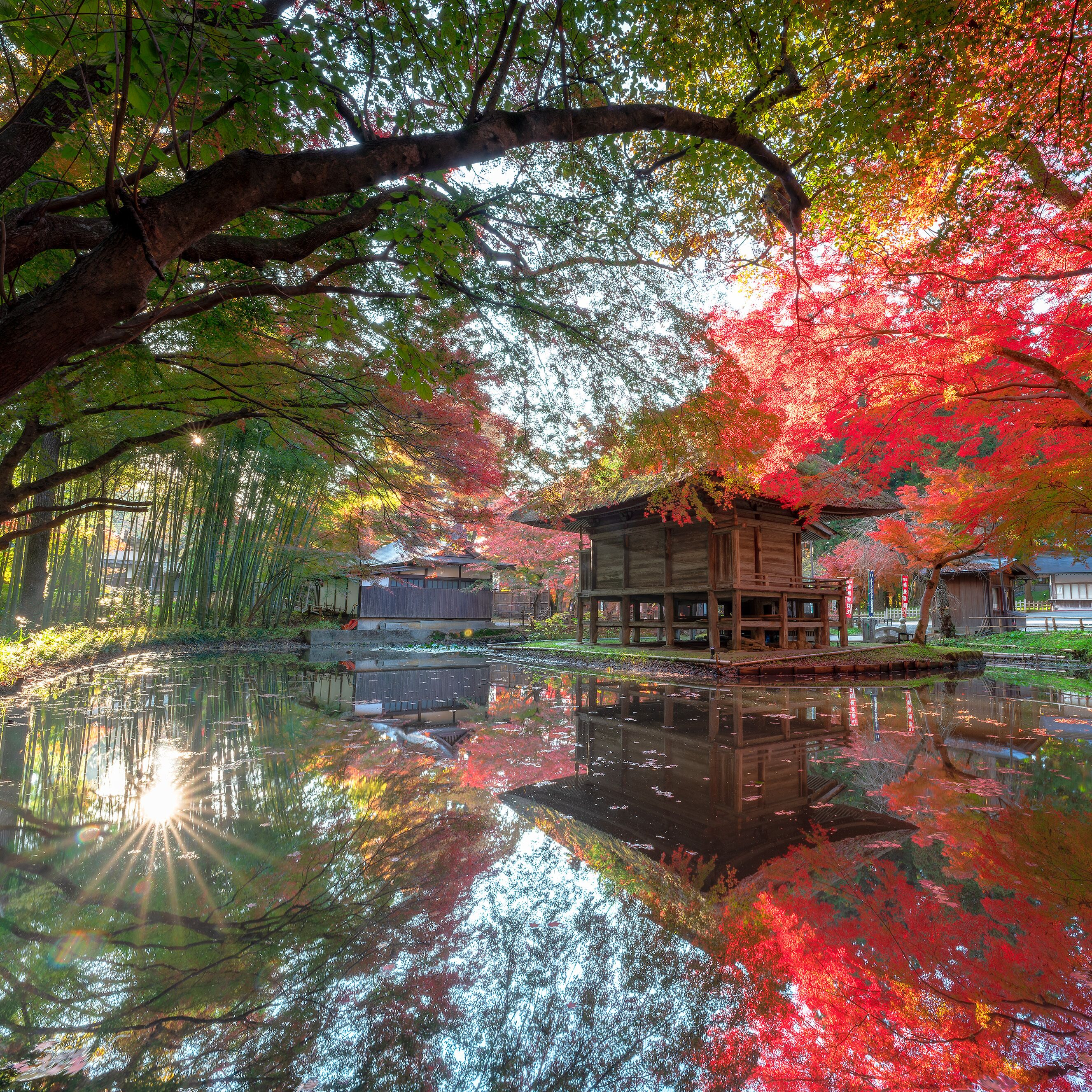 A traditional wooden structure by a reflective water body, surrounded by trees with autumn foliage.