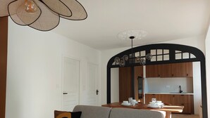 En moderne stue med en grå sofa, to mønstrede hynder, et spisebord med hvide tallerkener og kopper, samt et køkkenområde med træskabe og en sort buegang.