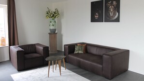A modern living room with a brown leather sofa, a matching armchair, a small round table, and a vase with flowers on a wooden stand. - Trompstraat (Eindhoven)