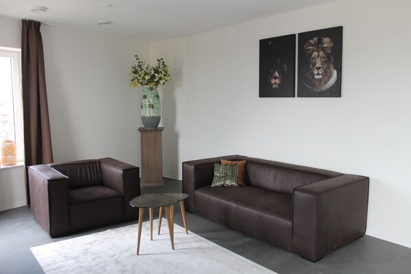 A modern living room with a brown leather sofa, a matching armchair, a small round table, and a vase with flowers on a wooden stand. - Trompstraat (Eindhoven)