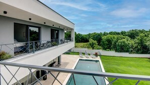 Una casa moderna de dos pisos con balcón, piscina y un paisaje verde exuberante al fondo.