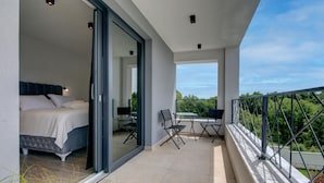 Un dormitorio moderno con una cama grande, un ventanal corredero que da a un balcón con mesa y sillas, y vistas a zonas verdes.