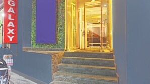 En hotellentrén med en skylt som visar texten ”HOTEL GALAXY”, en trappa som leder upp till entrén och en glasdörr.
