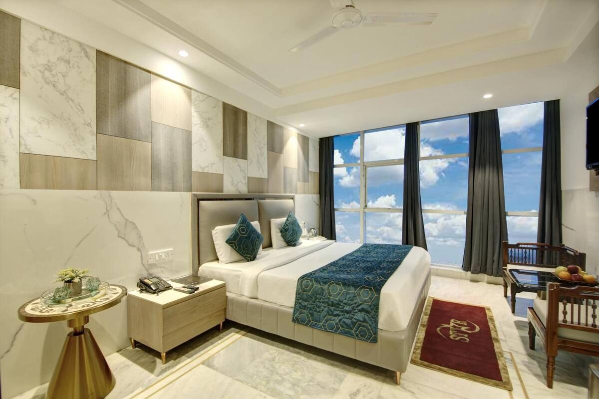 A modern bedroom with a large bed, a bedside table with a lamp, a round side table, and a view of the sky through large windows.