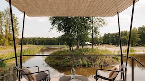 Een overdekt terras met twee stoelen en een tafel, met uitzicht op een rustige rivier en een bebost gebied.