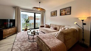 Un salon confortable avec un canapé, une table basse et un téléviseur. Deux tableaux encadrés ornent le mur, et une plante est posée sur le rebord de la fenêtre.