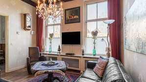 A living room with a sofa, a coffee table, a chandelier, a clock, a TV, and a window with curtains.
