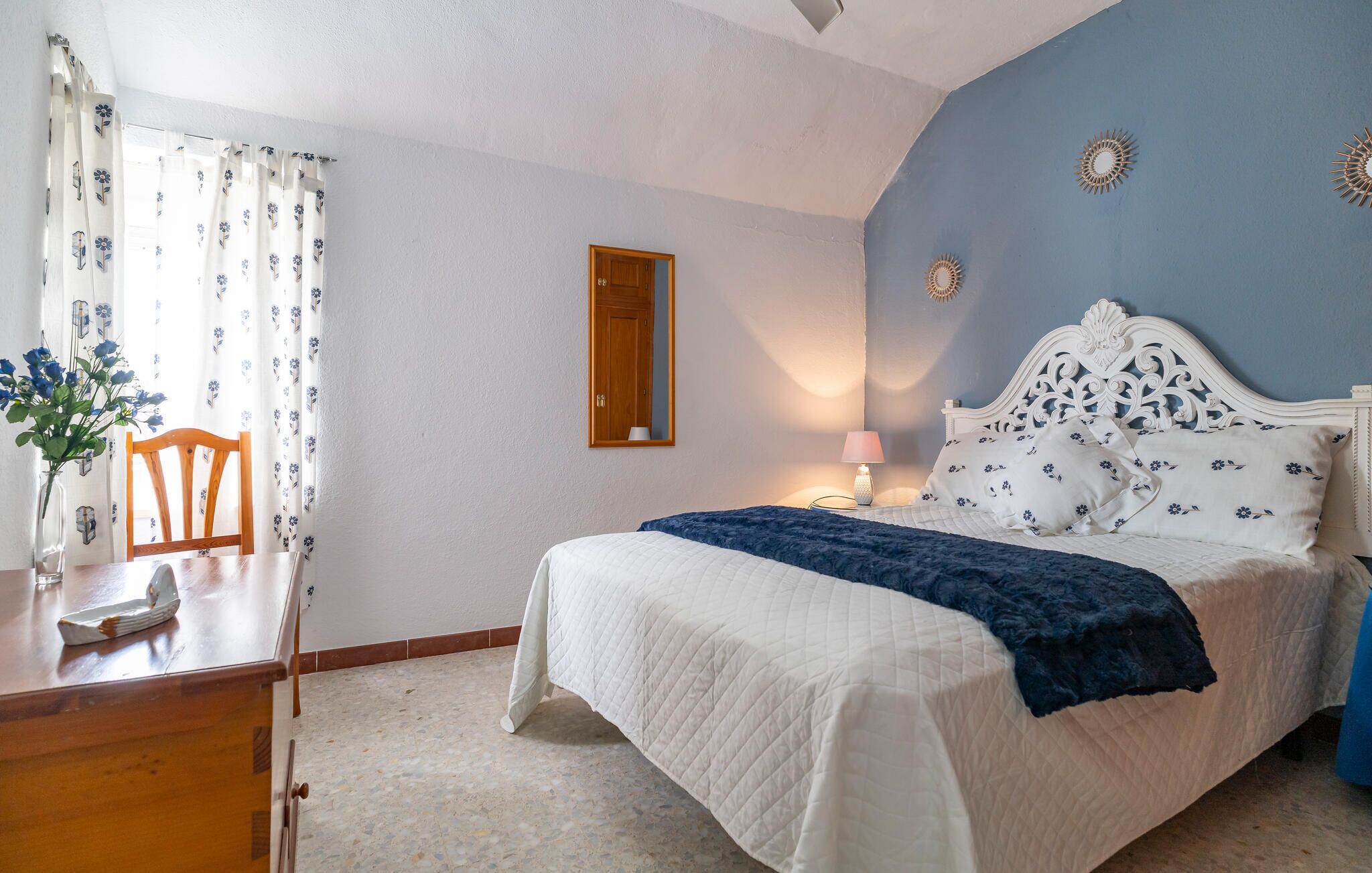 Un dormitorio con cama, una silla de madera, una mesa con un jarrón de flores y un espejo en la pared.