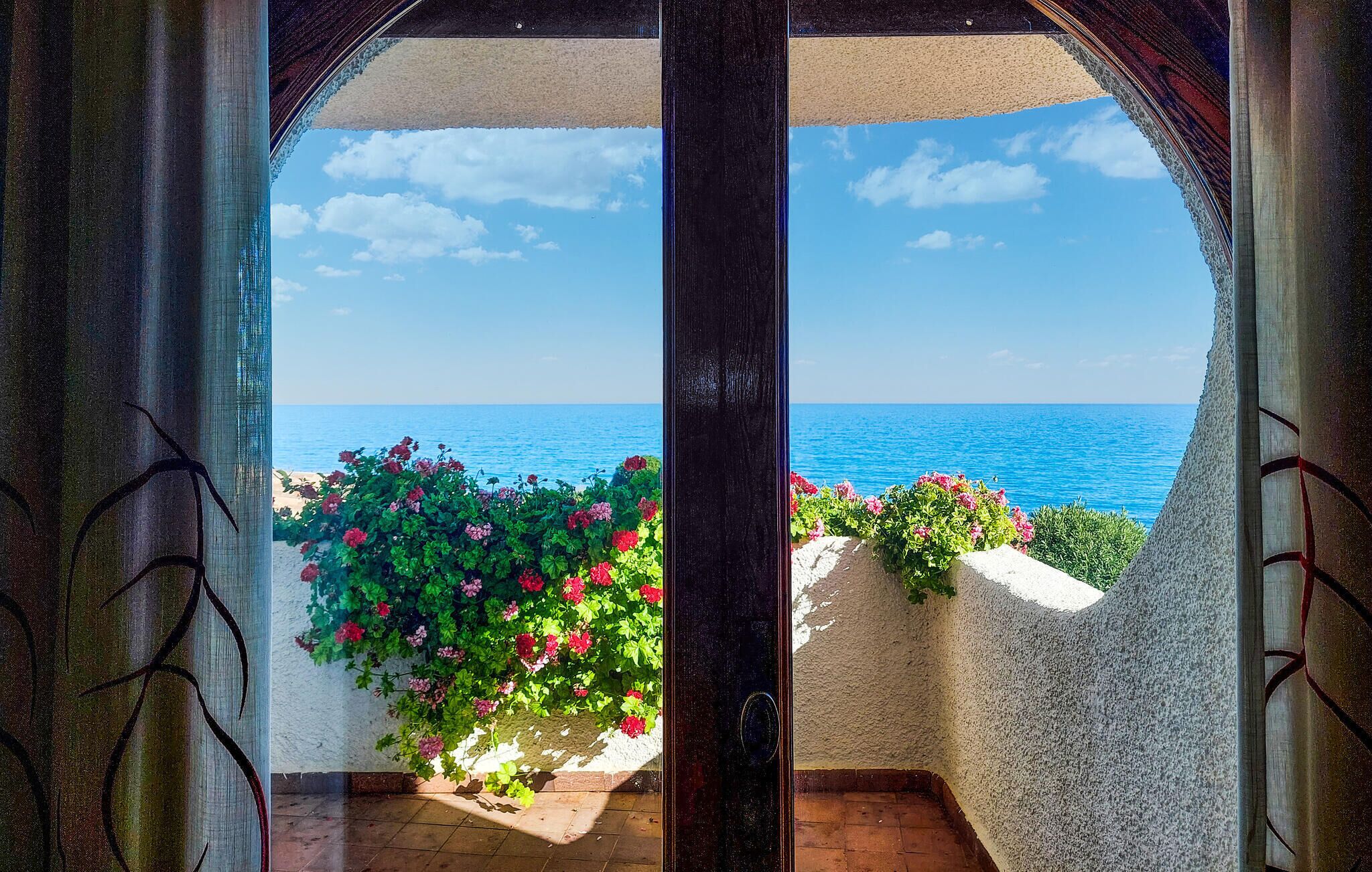 Uitzicht door een glazen deur op een balkon met uitzicht op zee, met rode bloemen en een heldere lucht.