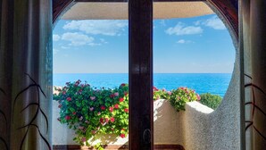 Blick durch eine Glastür auf einen Balkon mit Meerblick, mit roten Blumen und blauem Himmel.