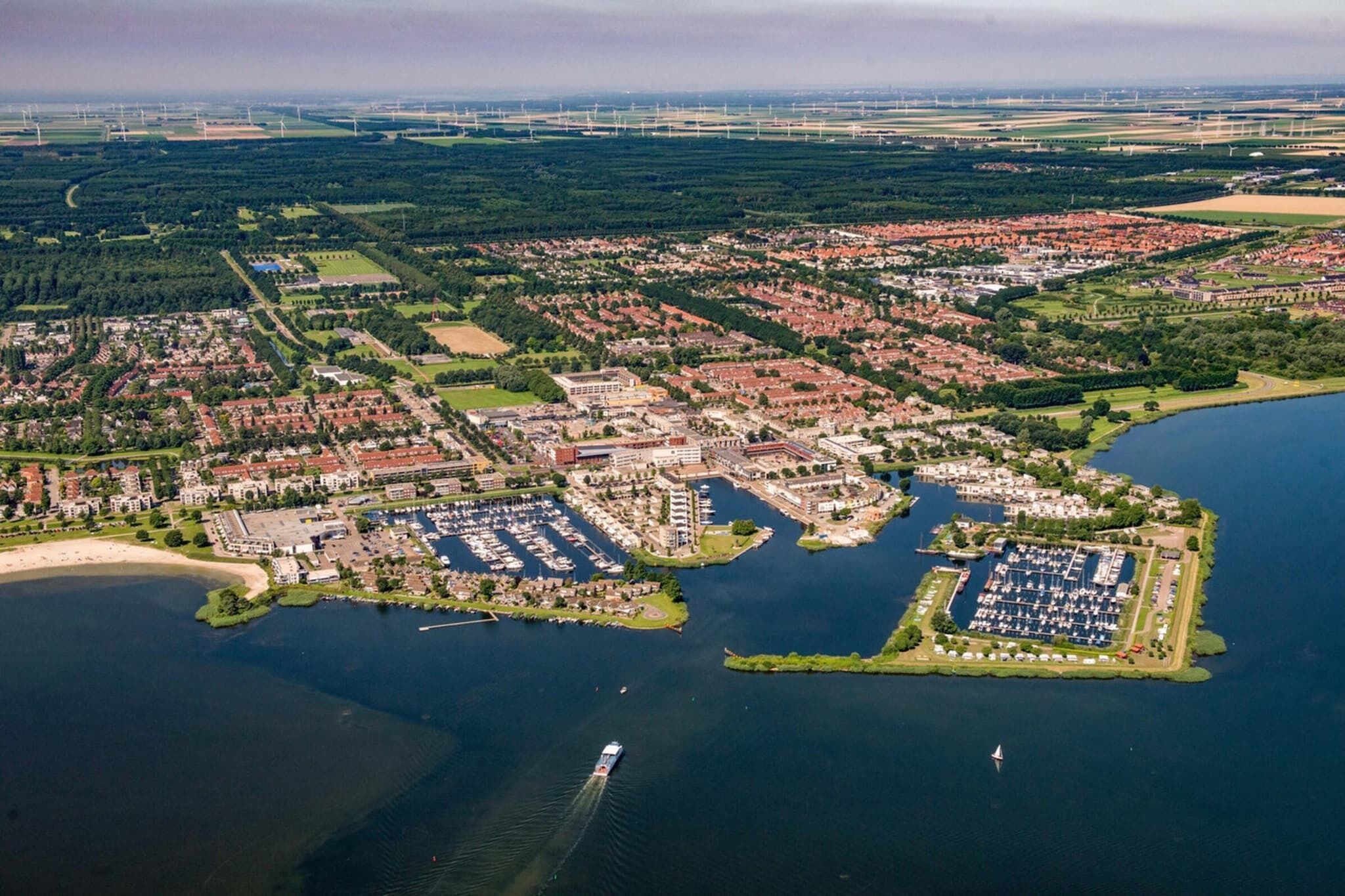 Luchtbeeld van een kuststad met een jachthaven, woonwijken en windturbines op de achtergrond.