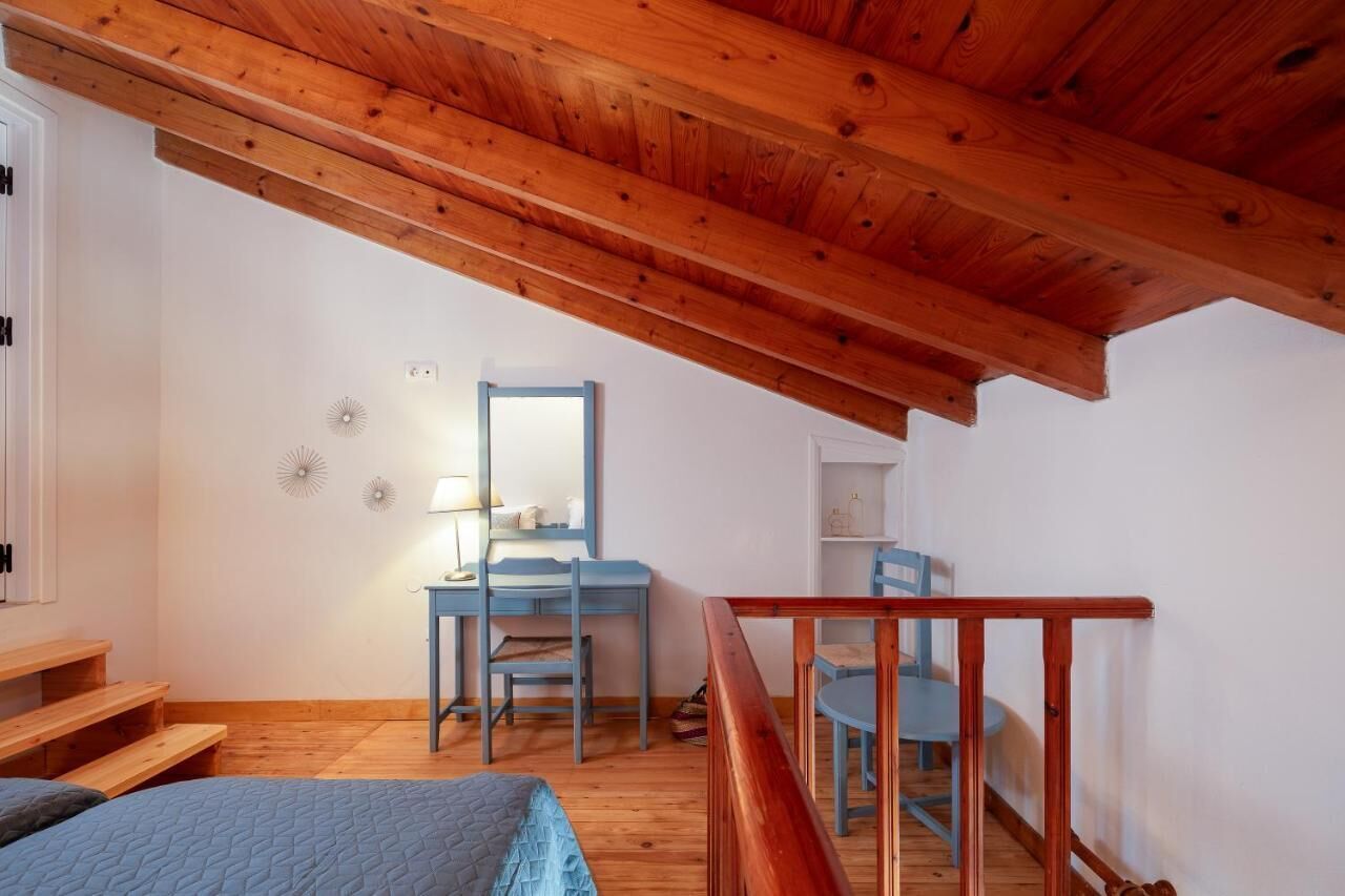 Quarto com teto de madeira, uma escrivaninha com cadeira, uma cama e uma escada.