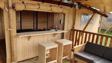 A wooden deck with a bench and two stools, a small table with a bottle and glasses, and a canopy overhead.