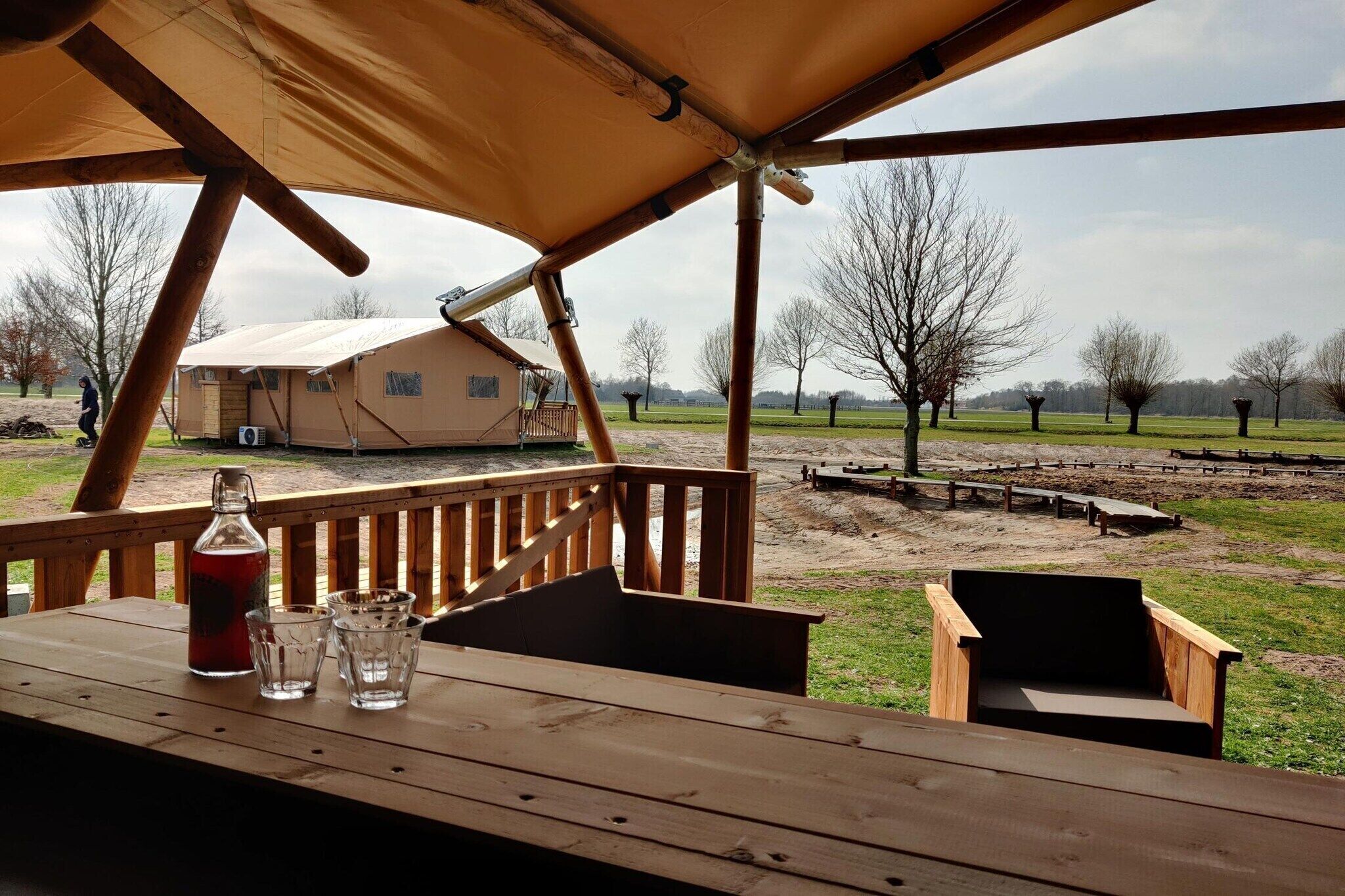 A wooden deck with a table, a bottle, and glasses. In the background, there is a house and a fenced area with trees.