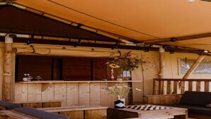 Een houten terras buiten, met zitplaatsen, een schaaktafel en een bar met flessen en bloemen.