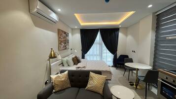 A modern hotel room with a bed, a sofa, a dining table, and a small kitchen area.