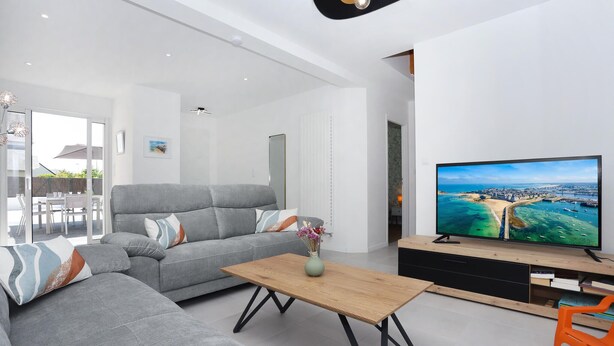 Un salón moderno con un sofá gris, una mesa de centro de madera y un televisor de pantalla plana mostrando una escena costera.