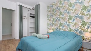 Un dormitorio con colcha color turquesa, un armario con perchero y un papel tapiz con estampado de hojas tropicales.