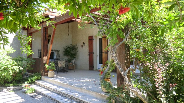 A covered outdoor area with a stone pathway leading to a wooden bench and a table, surrounded by greenery and potted plants. - Stunning home in Vedène (Vedène)