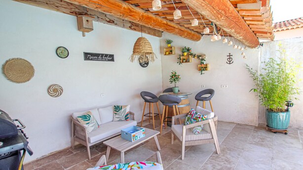 A cozy outdoor seating area with a wooden table, two cushioned chairs, and a wicker sofa. There are potted plants and wall decorations, including a sign that reads "Maison du bonheur." - Villa contemporaine Provençale (Mormoiron)