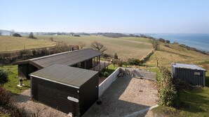 Een modern gebouw met een plat dak en een klein terras, omgeven door open velden en een waterpartij.