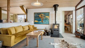 Een gezellige woonkamer met een bank, een houten tafel, een schilderij, een open haard en een groot raam.