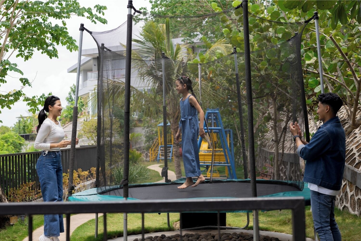 A trampoline in a garden with a safety net, surrounded by greenery and a playground structure.