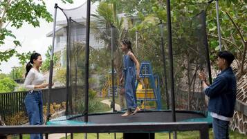 A trampoline in a garden with a safety net, surrounded by greenery and a playground structure.