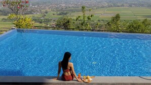Una mujer sentada junto a una piscina con una bandeja de comida, con vistas a un paisaje pintoresco.