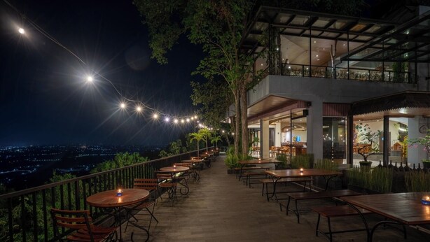Un espacio de comedor al aire libre con mesas y sillas, luces colgantes y vistas de la ciudad por la noche.