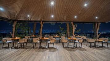 An outdoor dining area with round tables and wicker chairs, surrounded by wooden beams and large windows offering a city view at night.