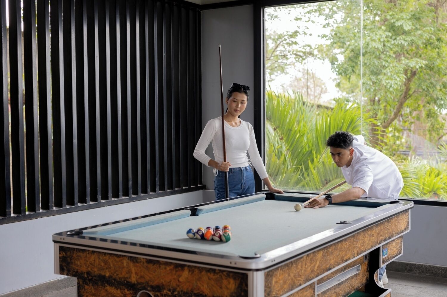 A pool table in a room with a large window, a black vertical blinds, and a view of greenery outside.