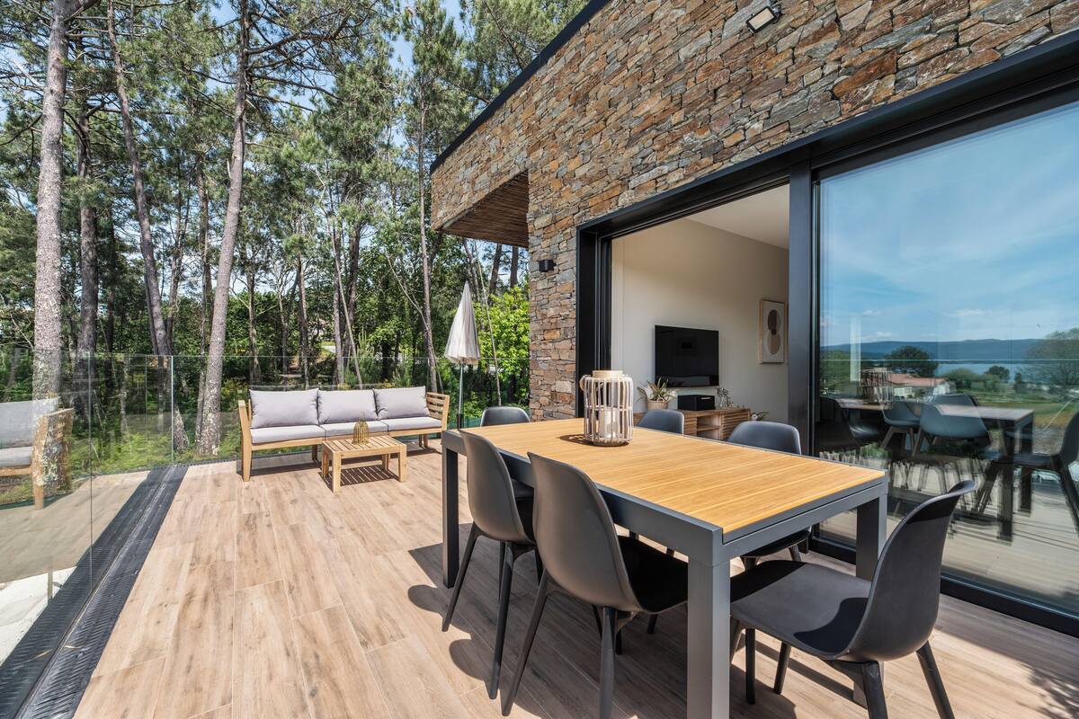 Un área de comedor exterior moderna con una mesa y sillas de madera, un sofá, y un amplio ventanal que da a una terraza con vistas al bosque y las colinas circundantes.