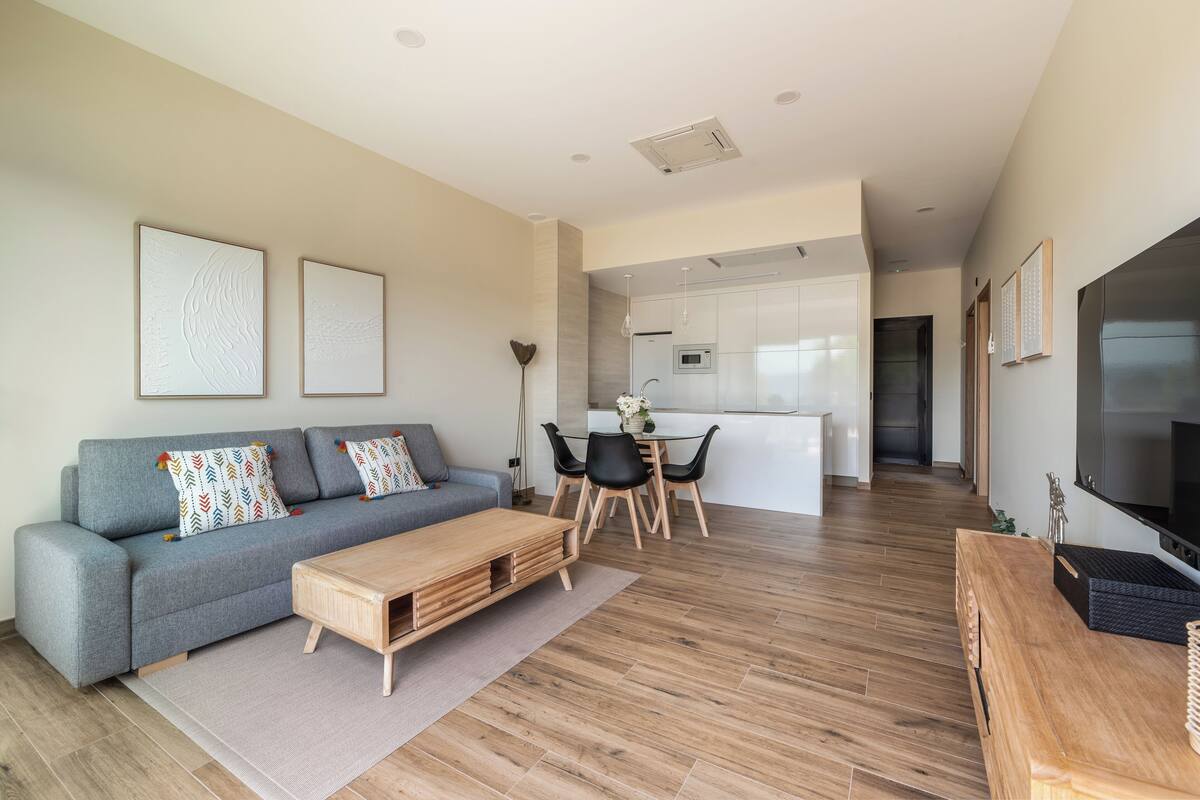 Una sala moderna con un sofá gris, una mesa de centro de madera y sillas negras. Se observa una cocina al fondo con armarios blancos y una mesa de comedor.