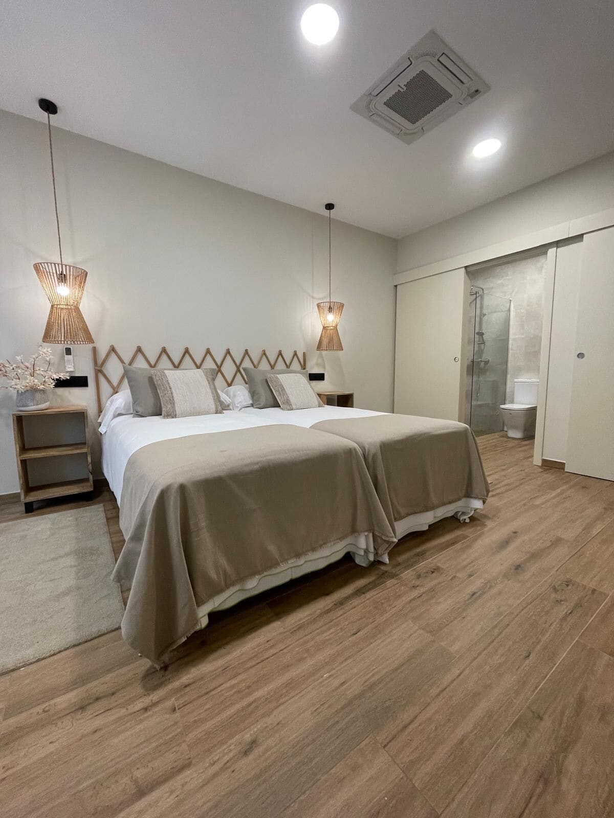 A bedroom with a bed, bedside tables, a nightstand, a hanging light fixture, a ceiling vent, and a bathroom with a toilet and shower.