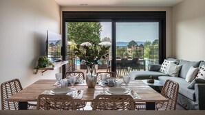 Ein Essbereich mit Holztisch, gedeckt für vier Personen, umgeben von Korbstühlen, einem großen Fenster mit Blick auf Grünflächen und einen Pool, sowie ein graues Sofa mit Kissen.