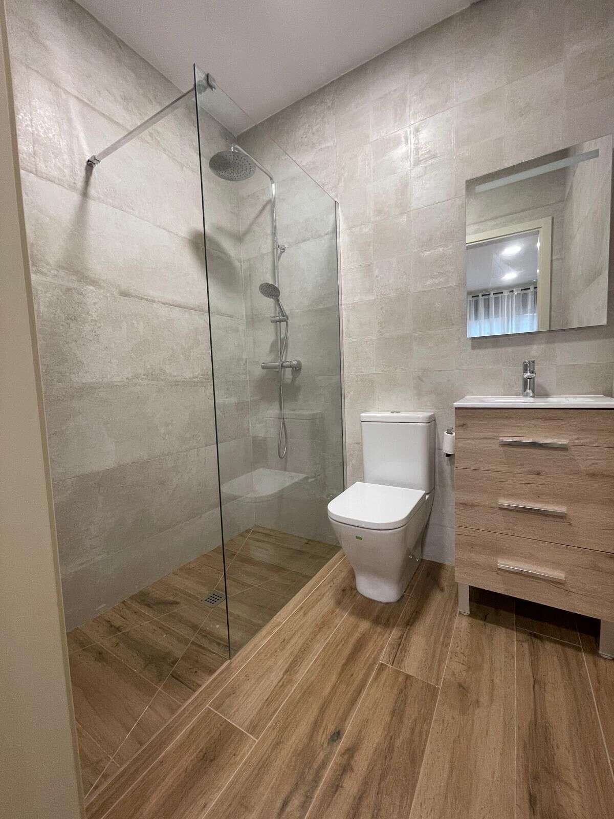 Un baño moderno con ducha envuelta en vidrio, inodoro blanco y tocador de madera con cajones.